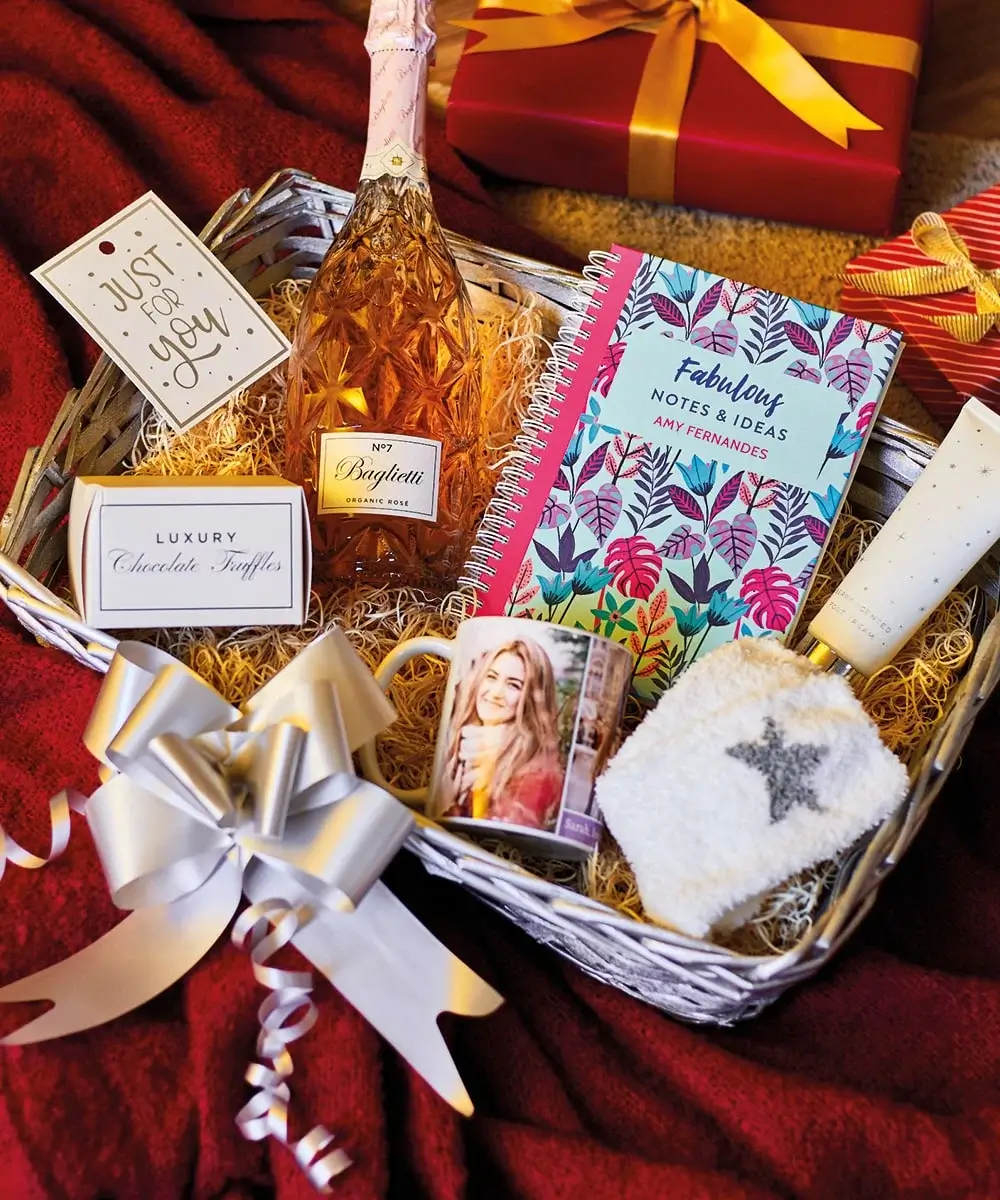 Christmas gift basket for her with a bottle of 'Brigatti No7' sparkling wine, a 'Fabulous Notes & Ideas' notebook, luxury truffles, lotion, a photo mug, and starry fuzzy socks, set on red fabric with a wrapped gift in the background.