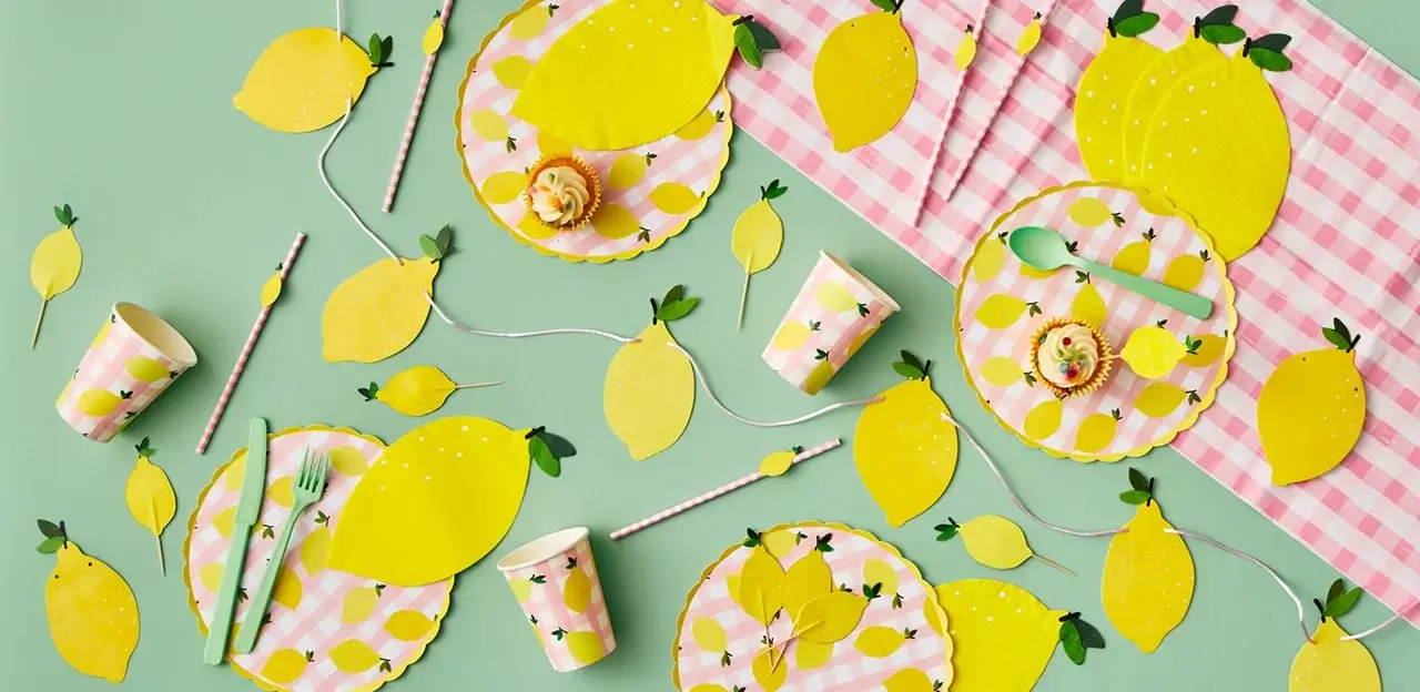 'The image shows a lemon-themed table setting with paper plates, cups, napkins, and utensils. The plates have a pink gingham pattern with yellow lemons around the edges. There are also lemon-shaped cutouts scattered across the table, along with green plastic forks and spoons. Cupcakes topped with lemon decorations are placed on some of the plates. The background is a light green surface.'