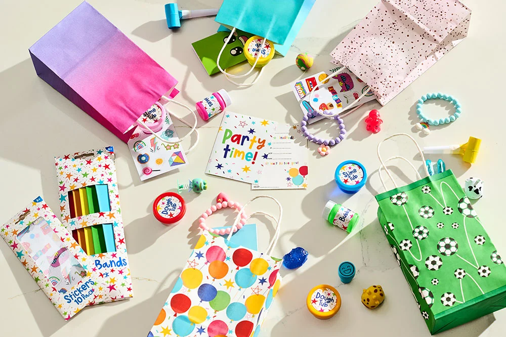 “A selection of children's party prizes and party bag fillers spread across a light background, including colourful gift bags, slime tubs, modelling dough, sticker sheets, crayons, novelty toys, bracelets, bubbles and party blowers. A ‘Party Time!’ invitation sits at the centre of the display, surrounded by assorted party game rewards and favour bag ideas.