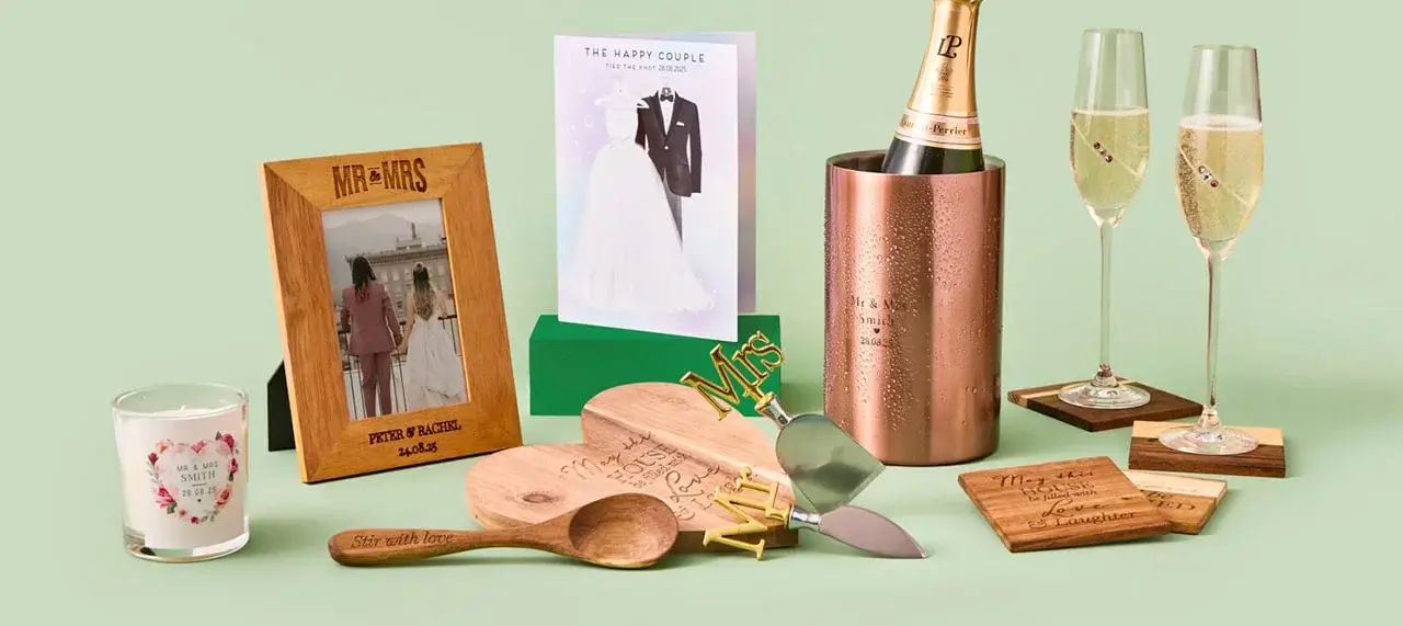 'A collection of wedding-themed items arranged on a light green background, including a framed photo, a candle, wooden spoons, a bottle of champagne in a copper cooler, two champagne glasses on coasters, a card with an illustration of a bride and groom, engraved cutting boards, and cake serving utensils with gold-colored 'Mr & Mrs' toppers.'