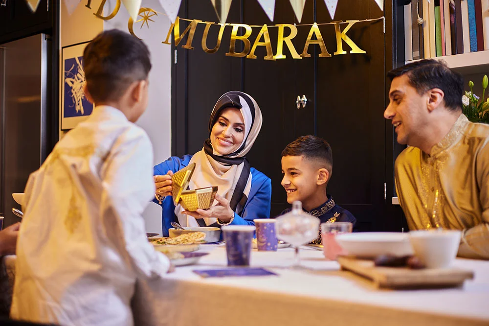 AA family gathered around a decorated table for an Eid meal, with food, drinks, and festive tableware, beneath a gold ‘Eid Mubarak’ banner.