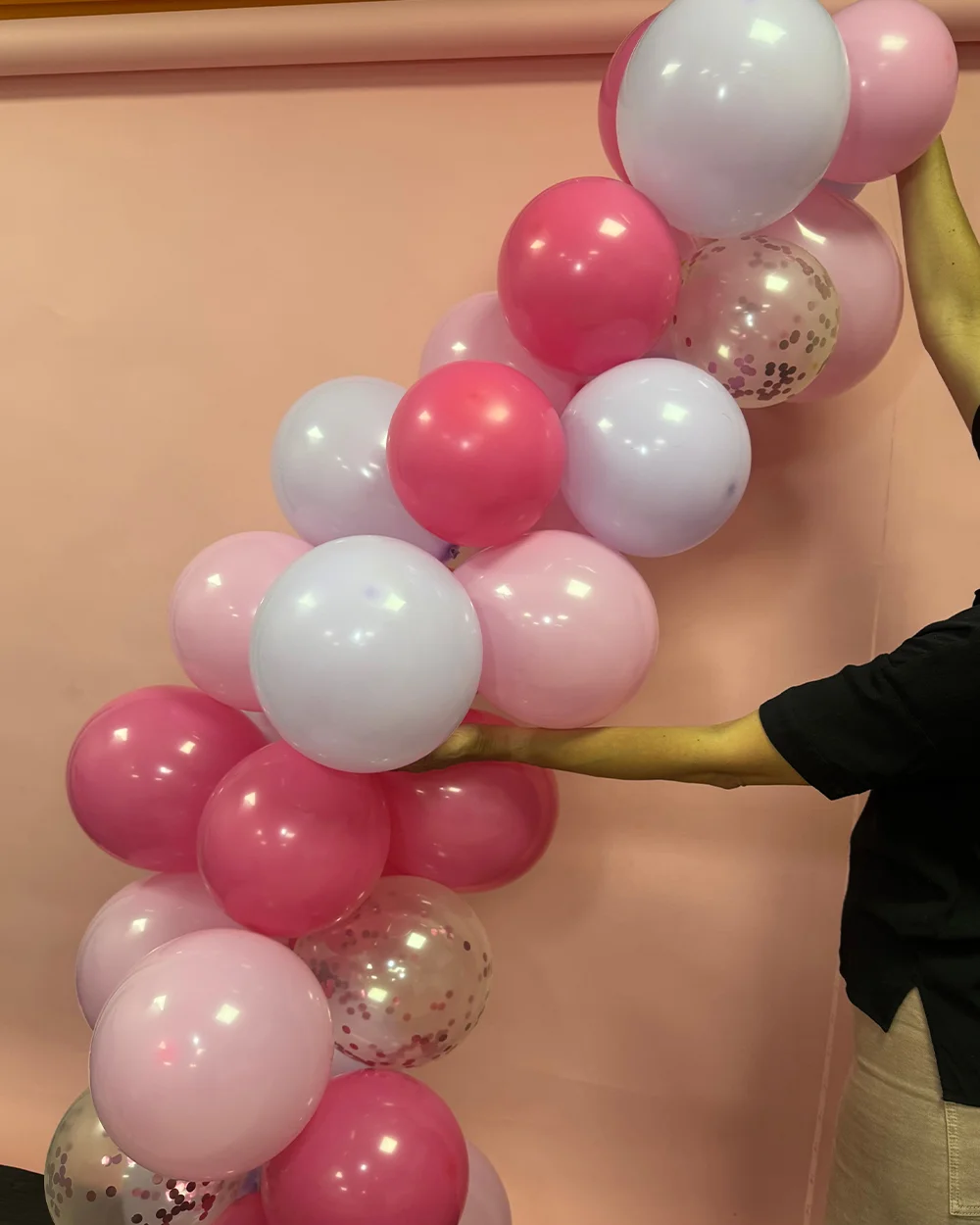 A partially assembled balloon arch made from clusters of pink, pale pink, white and clear confetti-filled balloons, arranged in a curved shape against a pastel pink backdrop. A person’s hands are shown positioning and shaping the balloon clusters, demonstrating how to build and structure a balloon arch decoration. A partially assembled balloon arch made from clusters of pink, pale pink, white and clear confetti-filled balloons, arranged in a curved shape against a pastel pink backdrop. A person’s hands are shown positioning and shaping the balloon clusters, demonstrating how to build and structure a balloon arch decoration.
