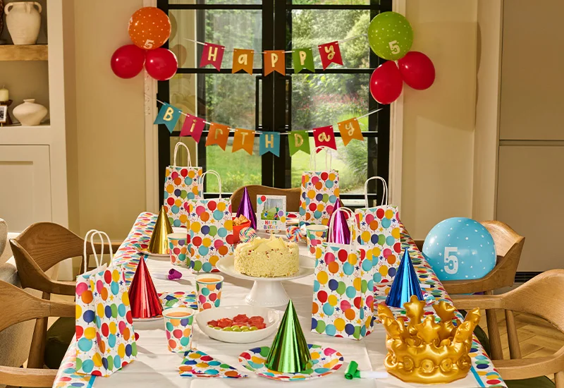 A colourful children's birthday party table decorated with balloon-print partyware, including matching gift bags, plates, cups and napkins. A birthday cake topped with a ‘Happy Birthday’ decoration sits at the centre of the table, surrounded by party hats, snacks and drinks. Above the table, a multicoloured ‘Happy Birthday’ bunting banner and clusters of balloons decorate the window, while a gold crown decoration and blue number 5 balloon complete the festive birthday setup.