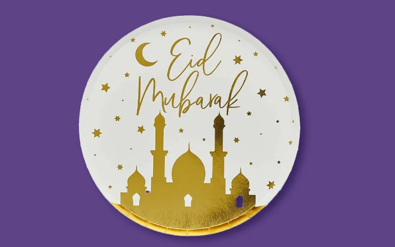 A round white plate decorated with a gold mosque silhouette, crescent moon and stars, featuring the text ‘Eid Mubarak,’ shown against a purple background.