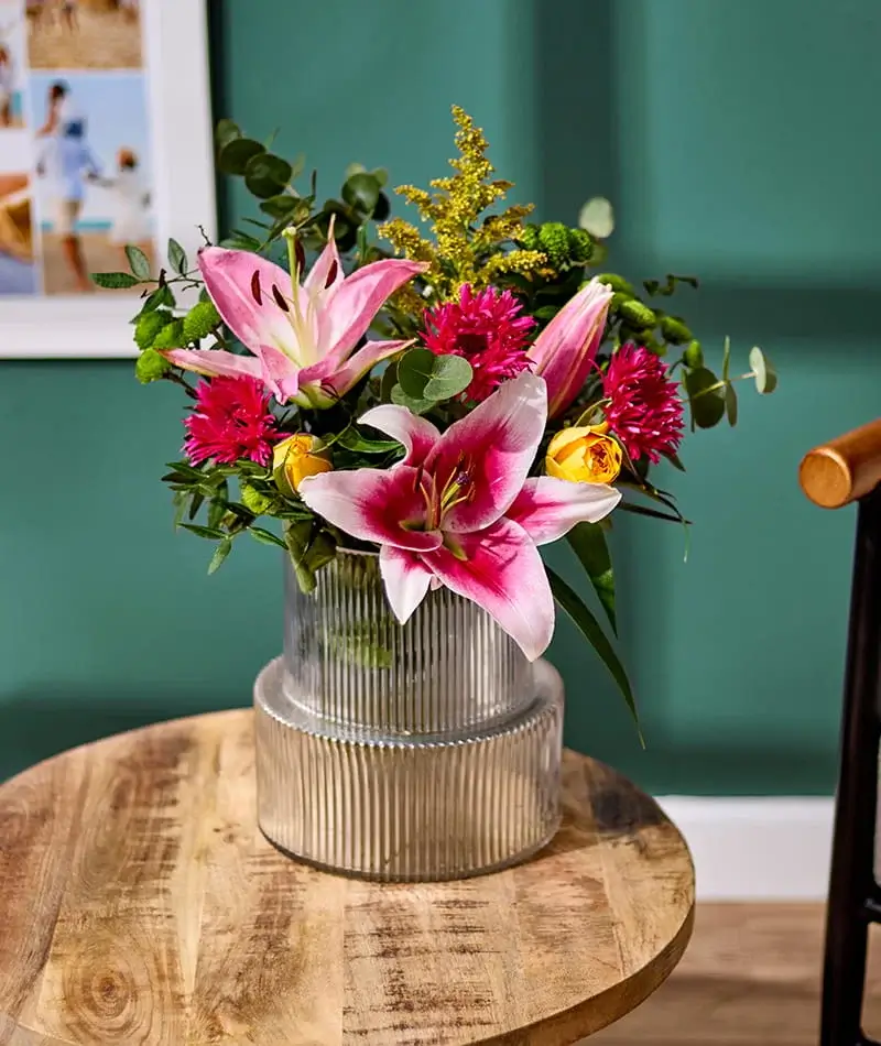 Floral bouquet with pink lilies and mixed flowers arranged in a ribbed glass vase on a wooden table against a green wall
