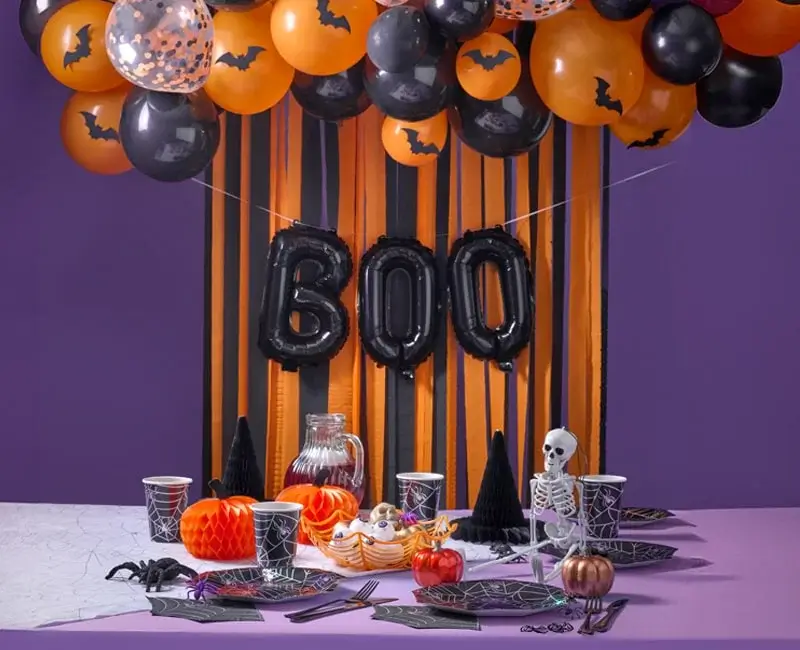 Halloween-themed table setting with black and orange decorations, including 'BOO' balloons, spiderweb-patterned tableware, a red drink pitcher, pumpkins, a skeleton figurine, and spooky accents.