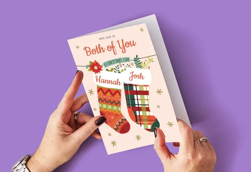 A person holding a Christmas card with two festive stockings labeled Hannah and Josh, surrounded by gold stars, against a purple background.