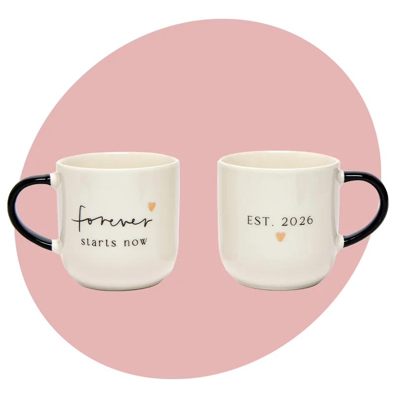 Two cream ceramic mugs with black handles, one reading ‘Forever starts now’ and the other ‘EST. 2026’ with a small gold heart detail, displayed against a soft pink circular background. Two cream ceramic mugs with black handles, one reading ‘Forever starts now’ and the other ‘EST. 2026’ with a small gold heart detail, displayed against a soft pink circular background.