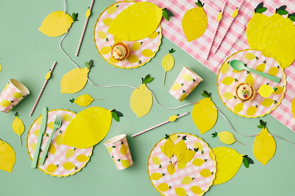 A lemon-themed party table setting arranged on a mint green background, featuring gingham pink-and-white plates decorated with lemon illustrations, matching paper cups, straws, napkins and cutlery. Lemon-shaped place settings, garlands and cupcake toppers are scattered throughout the display, alongside cupcakes and coordinating party decorations in yellow, pink and green. A lemon-themed party table setting arranged on a mint green background, featuring gingham pink-and-white plates decorated with lemon illustrations, matching paper cups, straws, napkins and cutlery. Lemon-shaped place settings, garlands and cupcake toppers are scattered throughout the display, alongside cupcakes and coordinating party decorations in yellow, pink and green.