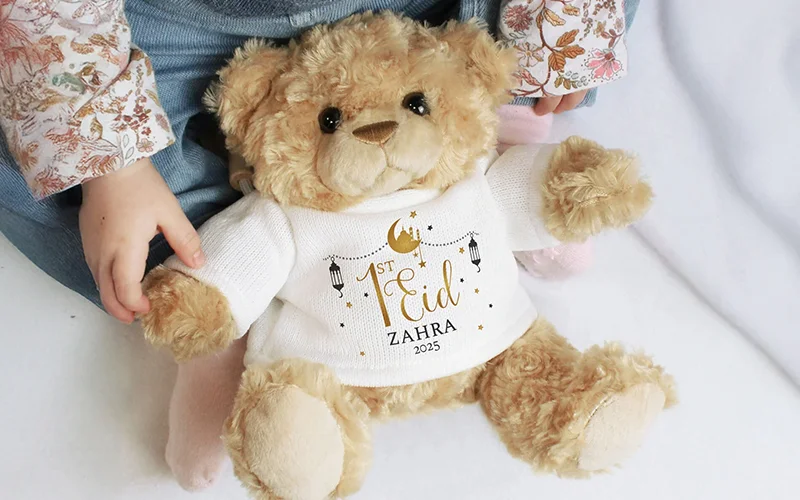 A soft plush teddy bear wearing a white jumper printed with gold crescent moons, stars and lanterns, personalised with the text ‘1st Eid Zahra 2025.’ A child’s hands rest beside the bear.