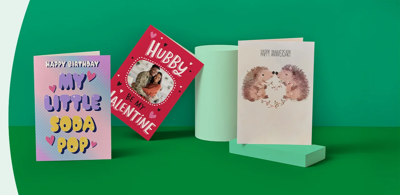 Three greeting cards on a green background: one with dogs saying 'Happy Wedding day! EMMA & JOHN', one with tomatoes saying 'On our Anniversary I love you from my head to-ma-toes', and one with a cow print saying 'HOLY COW! IT'S YOUR BIRTHDAY