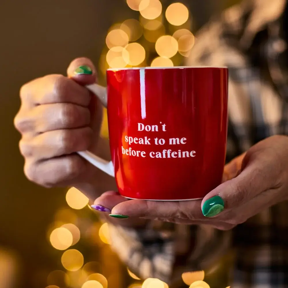 Person holding a red coffee mug with the text Don’t speak to me before caffeine, with festive lights blurred in the background