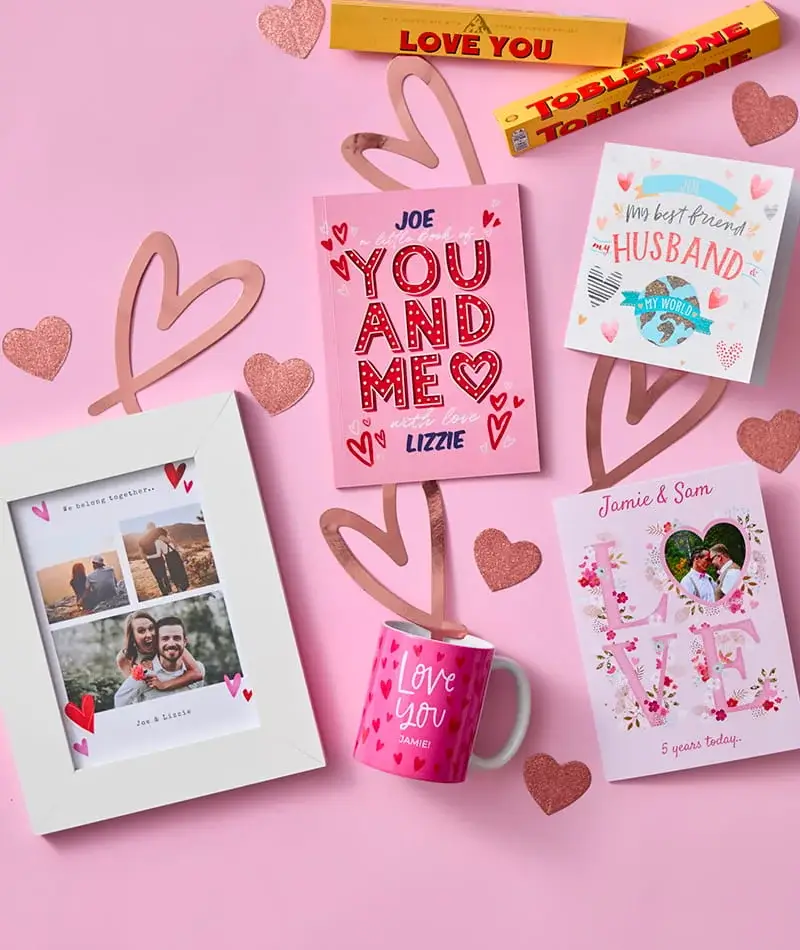 Anniversary gift collection featuring personalised cards, a photo frame, a personalised mug, chocolates, and romantic keepsakes on a pink background.