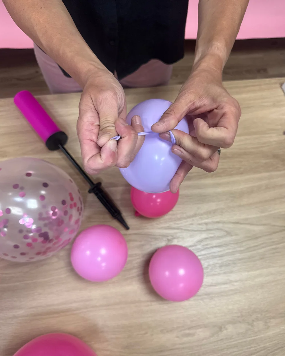 A step-by-step balloon arch assembly scene showing a pair of hands tying together inflated pink and lilac balloons on a wooden tabletop. Nearby are additional pink balloons, a clear confetti-filled balloon, and a pink hand pump, demonstrating how to create balloon clusters for a balloon arch decoration. A step-by-step balloon arch assembly scene showing a pair of hands tying together inflated pink and lilac balloons on a wooden tabletop. Nearby are additional pink balloons, a clear confetti-filled balloon, and a pink hand pump, demonstrating how to create balloon clusters for a balloon arch decoration.