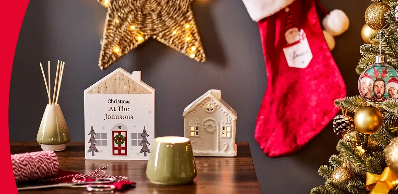 Festive Christmas scene with a decorated tree, red stocking, personalized house sign, ceramic house, lit star ornament, reed diffuser, candle holder, and wrapped gifts Festive Christmas scene with a decorated tree, red stocking, personalized house sign, ceramic house, lit star ornament, reed diffuser, candle holder, and wrapped gifts
