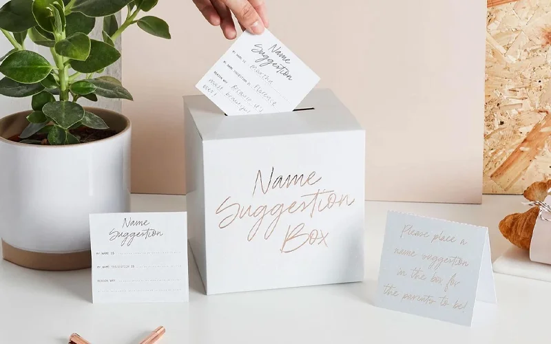 A white baby shower name suggestion box with gold script lettering reading ‘Name Suggestion Box’, shown with suggestion cards and a hand placing a completed card into the slot. Additional signs and cards are displayed beside a potted plant.