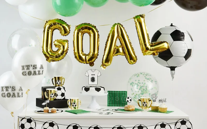 A football-themed party table decorated with green, white and gold balloons, featuring large gold foil letters spelling ‘GOAL,’ football-print tableware, trophy-shaped cups, football balloons and a football-themed cake displayed on a white cake stand. A football-themed party table decorated with green, white and gold balloons, featuring large gold foil letters spelling ‘GOAL,’ football-print tableware, trophy-shaped cups, football balloons and a football-themed cake displayed on a white cake stand.