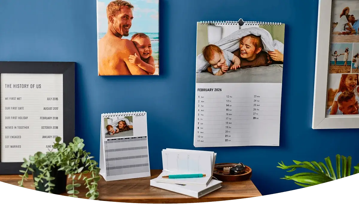 Personalised photo calendars and keepsake prints displayed on a desk with plants, notebooks, and framed family photographs against a blue wall. Personalised photo calendars and keepsake prints displayed on a desk with plants, notebooks, and framed family photographs against a blue wall.