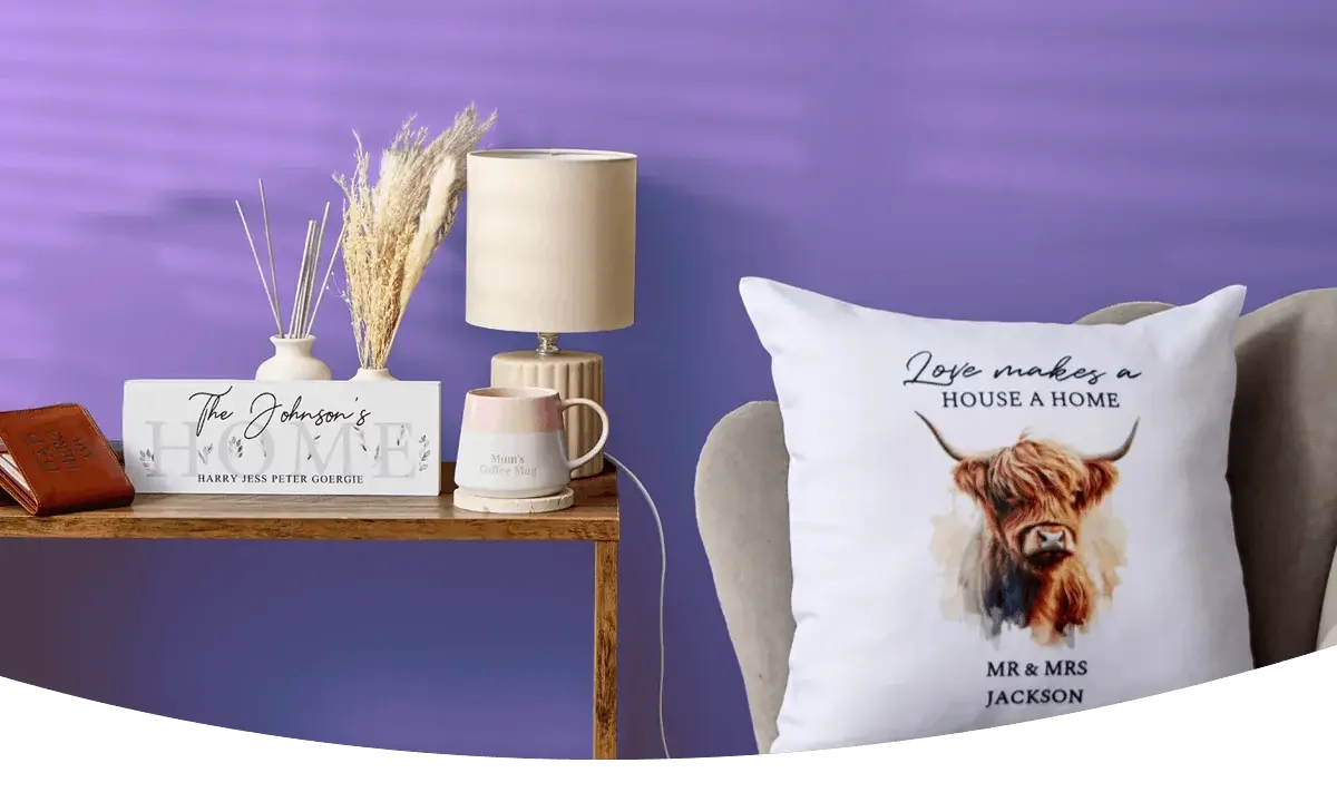 A cosy home display featuring personalised wall art, a wooden clock, a table lamp, framed prints, decorative accessories and a cushion with a highland cow design arranged against a dark blue wall.