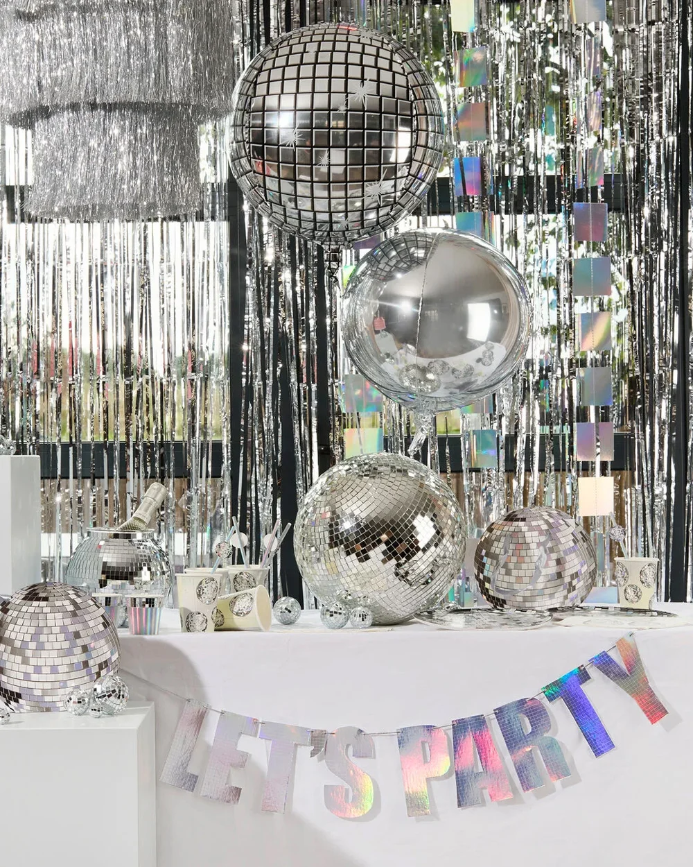 A disco-themed party display featuring mirrored disco ball decorations, silver tinsel curtains and iridescent hanging streamers. The table is decorated with reflective silver balloons, miniature disco balls, party cups and tableware, with a holographic banner reading ‘LET’S PARTY’ draped across the front, creating a sparkling silver celebration theme.