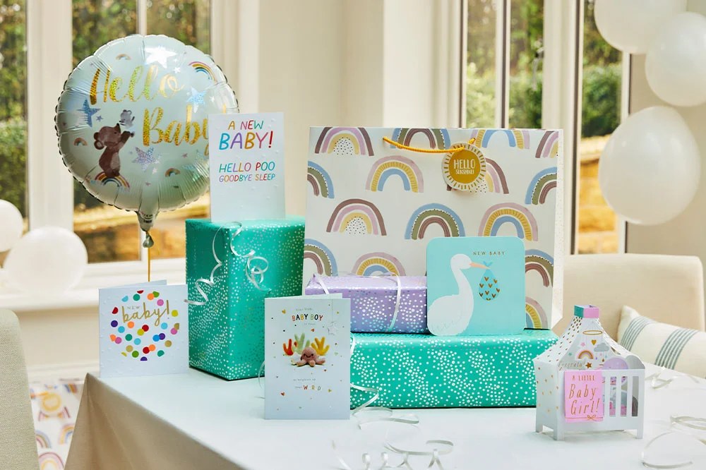 A baby shower gift table featuring wrapped presents, rainbow-patterned gift bags, baby-themed greeting cards and a ‘Hello Baby’ foil balloon. The display includes a lantern-style keepsake reading ‘A Little Baby Girl,’ alongside coordinated gift wrap, ribbons and baby celebration decorations arranged in a bright indoor setting.