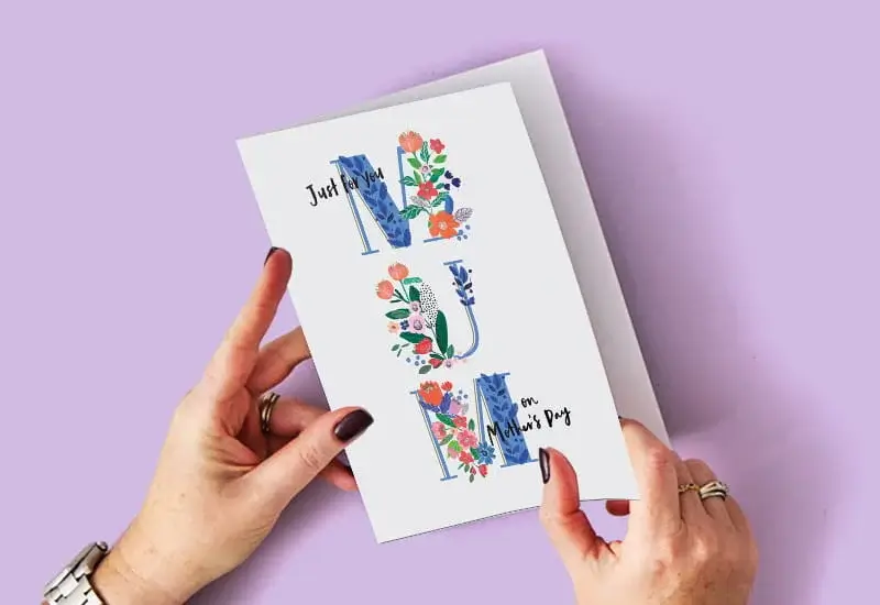 A person holding a floral Mother’s Day card with the word MUM illustrated in decorative blue letters filled with colorful flowers, set against a light purple background.