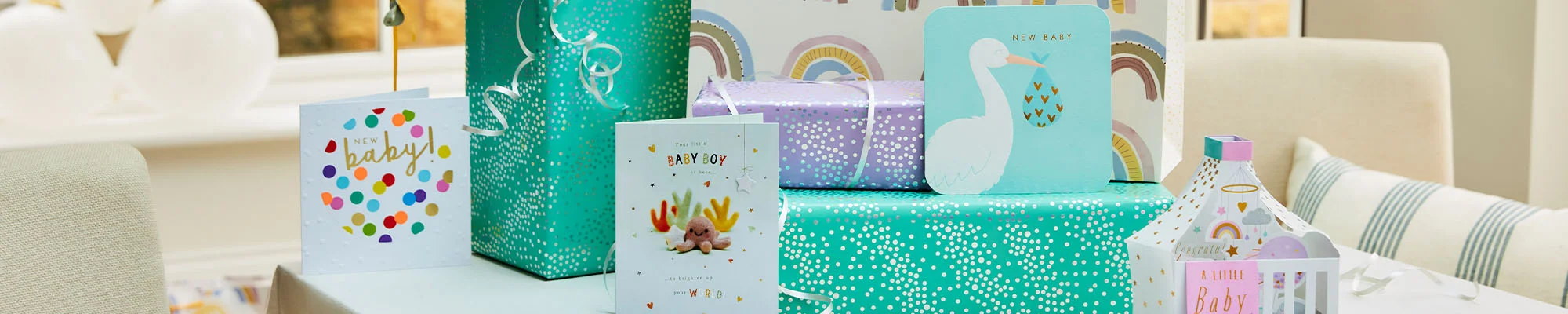 A baby gift display arranged on a table, featuring wrapped presents in teal and lilac gift wrap, baby-themed greeting cards, a large gift bag with a rainbow pattern, a ‘Hello Baby’ foil balloon, and a decorative lantern-style keepsake with baby-themed illustrations. White balloons and baby shower decorations complete the celebration display.