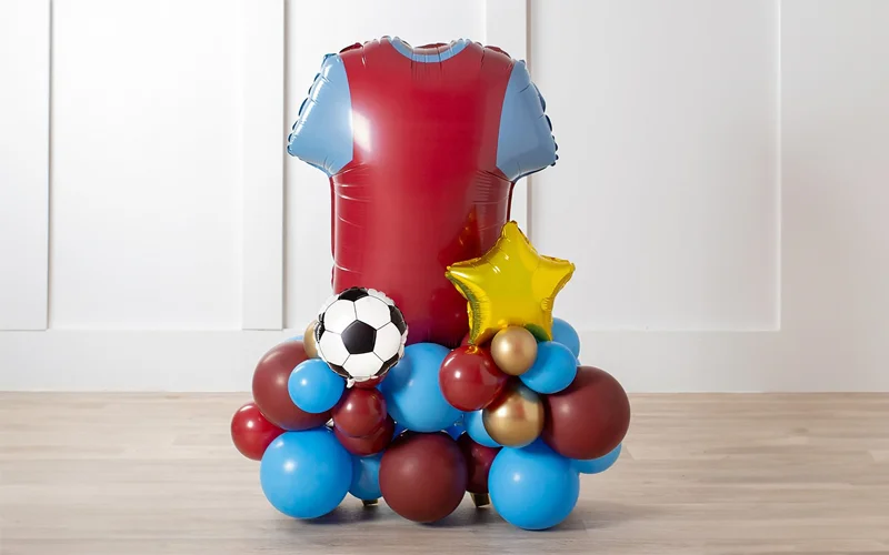 A football-themed balloon display featuring a large red shirt-shaped foil balloon mounted on a base of blue, burgundy and gold balloons, decorated with a football balloon and a gold star balloon, displayed against a white panelled wall. A football-themed balloon display featuring a large red shirt-shaped foil balloon mounted on a base of blue, burgundy and gold balloons, decorated with a football balloon and a gold star balloon, displayed against a white panelled wall.