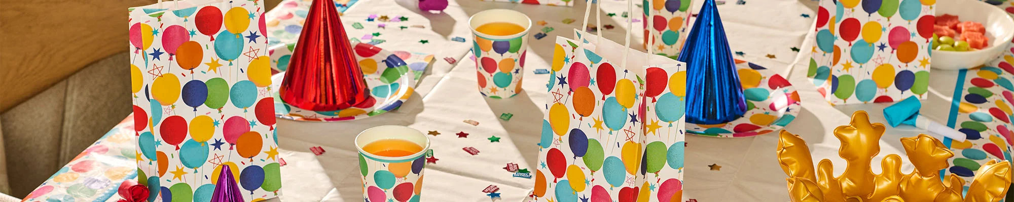 A colourful birthday party table setting featuring balloon-print gift bags, matching paper cups, plates and napkins arranged on a white tablecloth. Metallic party hats in red, blue and purple, scattered confetti and a gold crown decoration complete the bright birthday celebration display.