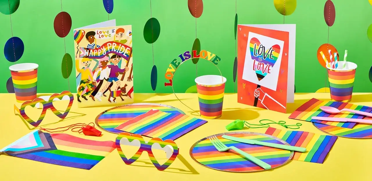 'The image shows a colorful Pride-themed party setup with rainbow decorations. There are two greeting cards, one with the text 'Love is Love' and another with 'Happy Pride'. The table is adorned with rainbow-striped plates, cups, napkins, and utensils. Heart-shaped glasses and a headband with the text 'Love is Love' are also present. The background features hanging circular decorations in various colors.'
