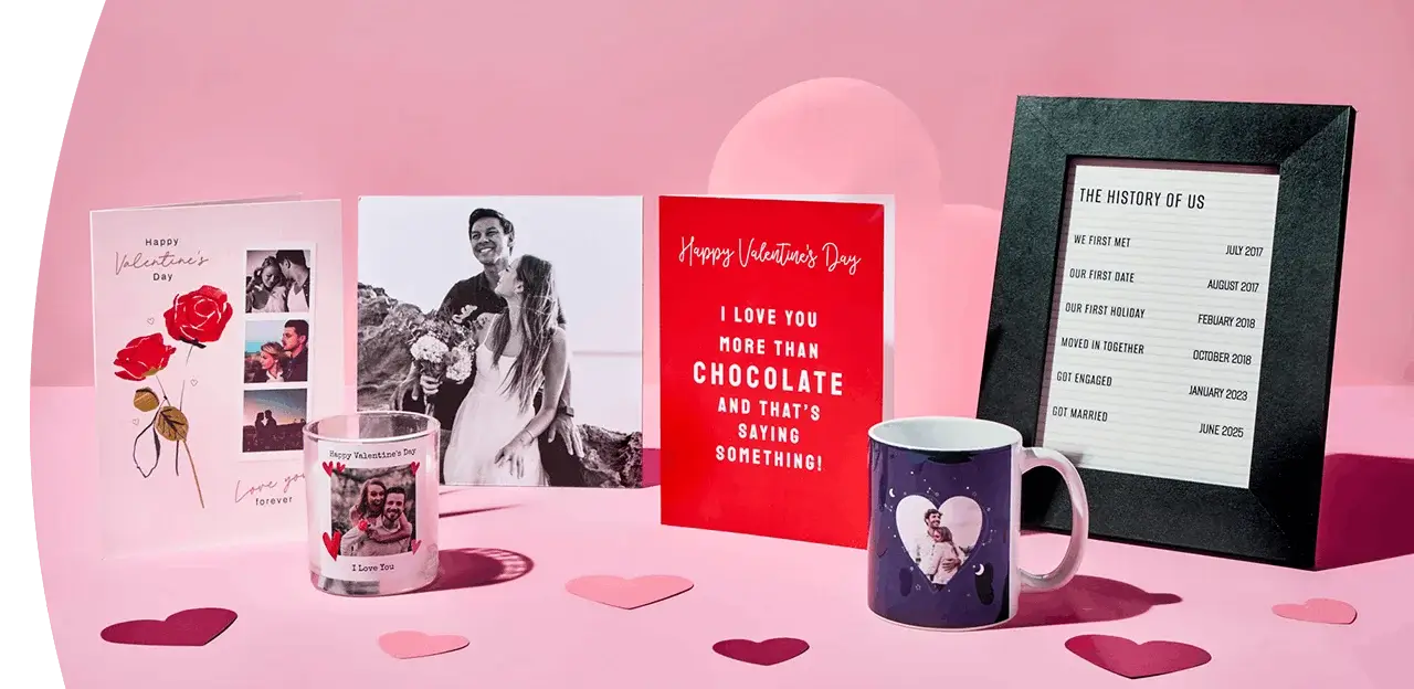 A display of Valentine’s gifts on a pink surface, including greeting cards, two photo mugs, heart‑shaped confetti, and a framed timeline titled The History of Us.