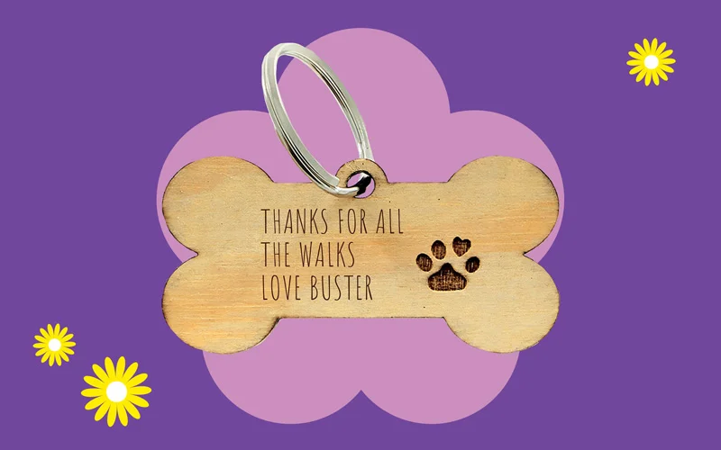 A wooden dog bone‑shaped keyring engraved with the message ‘Thanks for all the walks, love Buster,’ displayed against a purple background with a pink flower shape and small yellow daisies.
