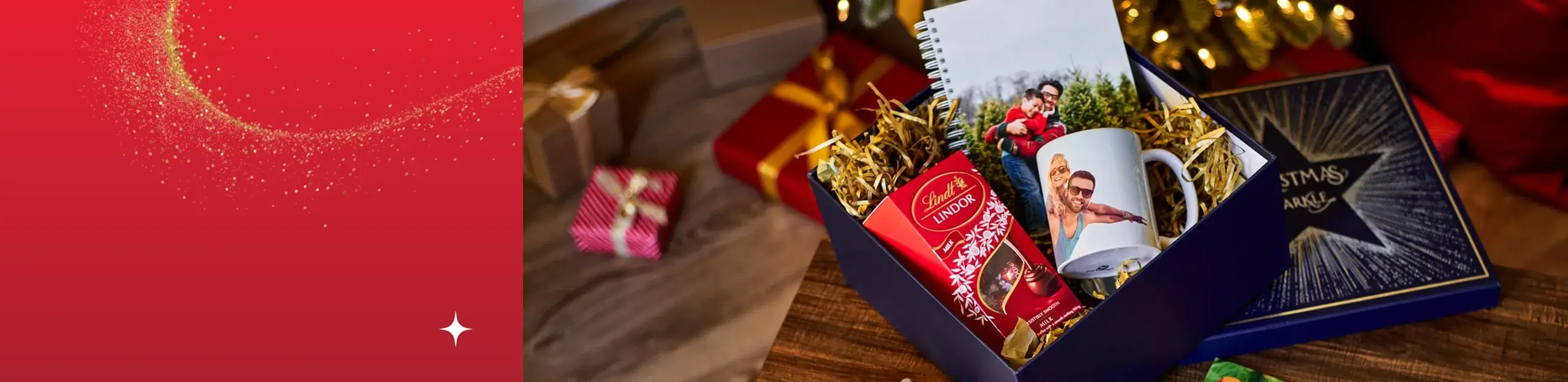Gift box containing Lindt chocolates, a custom photo mug, and a spiral notebook, placed on a wooden surface with wrapped presents and a Christmas tree in the background.