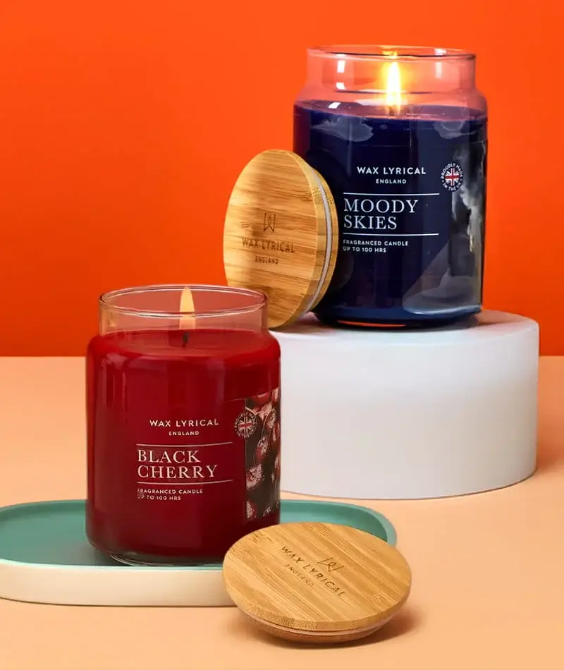 Two scented candles in glass jars with wooden lids, one black cherry and one moody skies, displayed on tiered stands against a bright orange background.