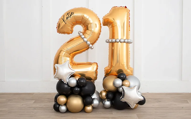 A 21st birthday balloon arrangement featuring large gold foil number balloons ‘2’ and ‘1’, personalised with the name ‘Katie’. The display is set on bases of black, gold and silver balloons, with silver star-shaped foil balloons and decorative balloon garlands creating a milestone birthday centrepiece. A 21st birthday balloon arrangement featuring large gold foil number balloons ‘2’ and ‘1’, personalised with the name ‘Katie’. The display is set on bases of black, gold and silver balloons, with silver star-shaped foil balloons and decorative balloon garlands creating a milestone birthday centrepiece.