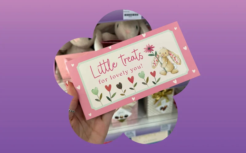 A gold heart-shaped Lindt Lindor chocolate box opened to reveal assorted chocolate truffles, displayed against a purple and pink gradient background with a flower silhouette.