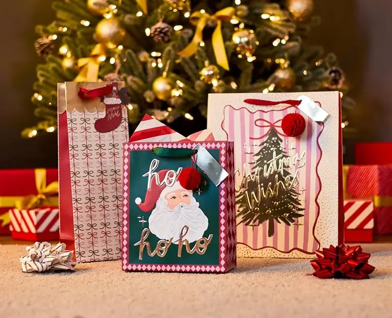 Christmas gift bags with festive designs, including Santa and a Christmas tree, placed on a carpet in front of a decorated tree with wrapped presents and bows