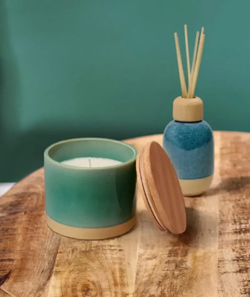 Ceramic candle and reed diffuser set with wooden lids displayed on a rustic wooden table against a teal background