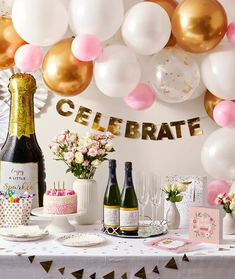 Celebration party table with champagne bottles, cake, flowers, greeting cards, and pink, white, and gold balloons with a celebrate banner