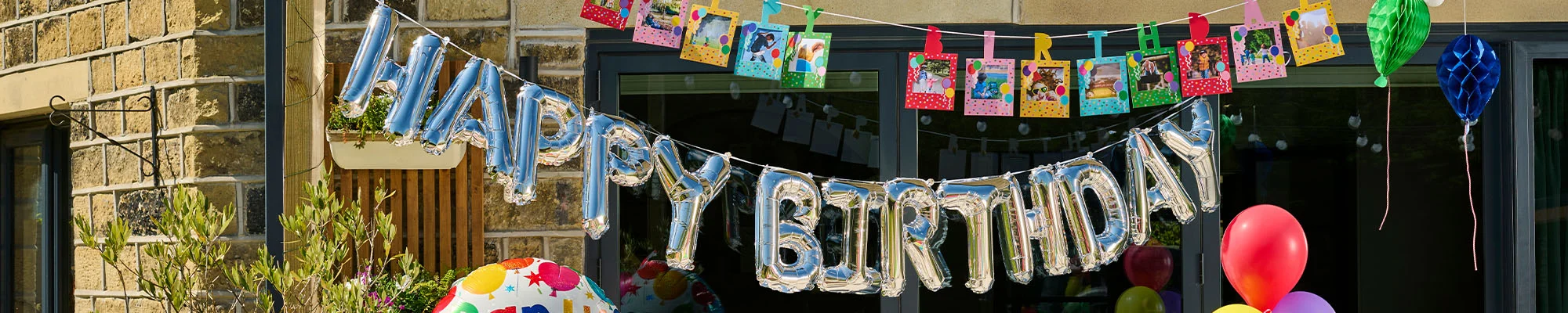 A silver foil ‘Happy Birthday’ balloon banner displayed across the front of a house, decorated with colourful birthday photo bunting showing snapshots clipped to a string above. Brightly coloured honeycomb decorations and balloons surround the display, creating a festive outdoor birthday celebration scene.