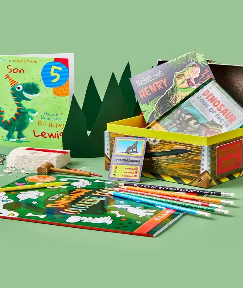 A collection of dinosaur-themed items arranged on a green surface. Includes a birthday card with a cartoon dinosaur and the text 'For a roarsome Son, Have a Dino-mite Birthday Lewis' and the number 5, a yellow box with books titled 'Dinosaur Book of Facts' and 'Henry', pencils, an activity book titled 'Dinosaurs', a small card labeled 'Tyrannosaurus', and a block of clay with excavation tools. A collection of dinosaur-themed items arranged on a green surface. Includes a birthday card with a cartoon dinosaur and the text 'For a roarsome Son, Have a Dino-mite Birthday Lewis' and the number 5, a yellow box with books titled 'Dinosaur Book of Facts' and 'Henry', pencils, an activity book titled 'Dinosaurs', a small card labeled 'Tyrannosaurus', and a block of clay with excavation tools.