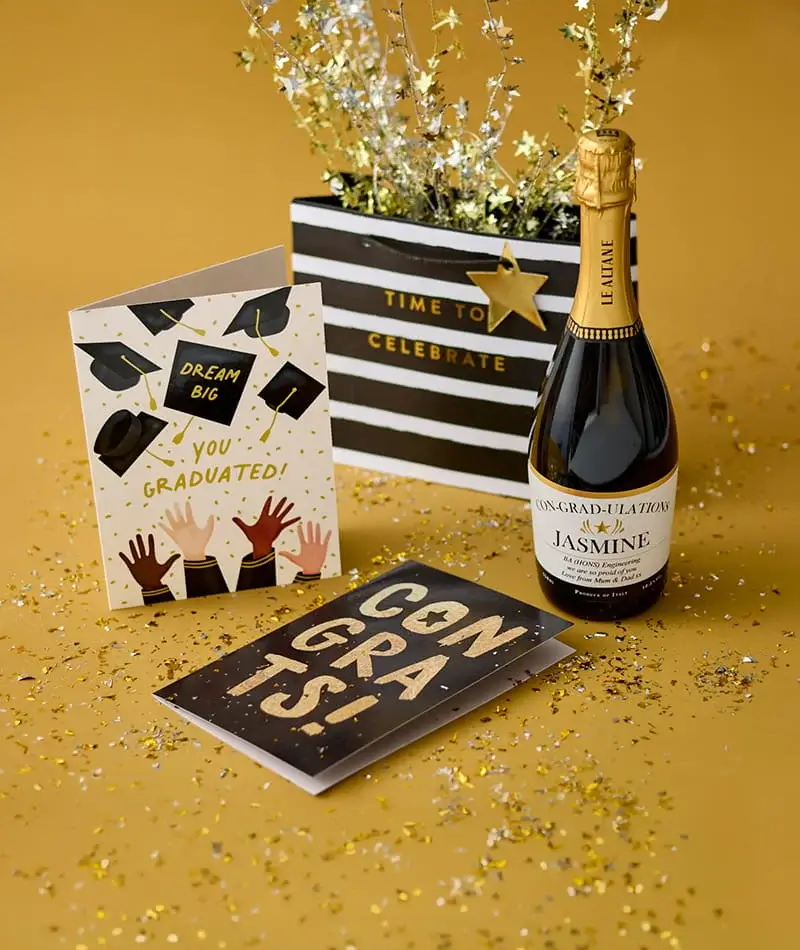 Graduation celebration display with congratulations cards, a personalised bottle of sparkling wine, gold confetti, and festive gold decorations.