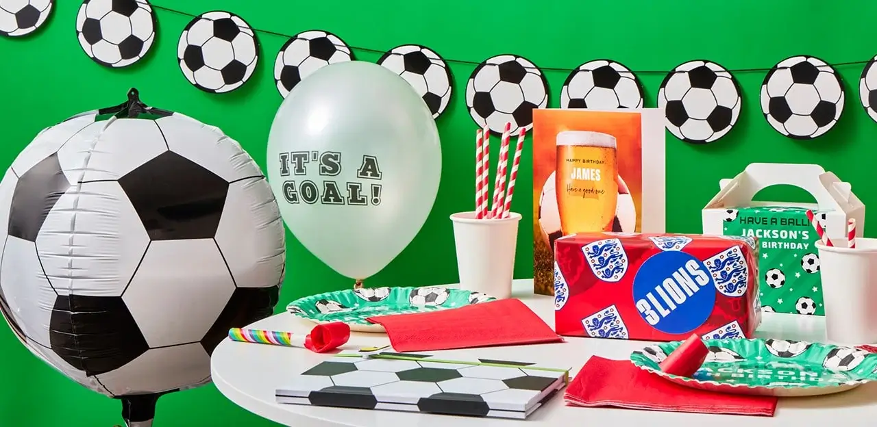 A soccer-themed birthday party setup with a large soccer ball balloon, a white balloon with the text 'IT'S A GOAL!', and various party supplies including plates, napkins, cups with striped straws, and a gift box. There is also a banner of soccer balls hanging in the background.