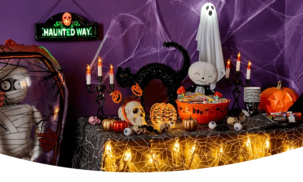 Halloween party table with spooky decorations, trick-or-treat sweets, glowing candles, pumpkins, cobwebs and a mummy figure against a purple backdrop.
