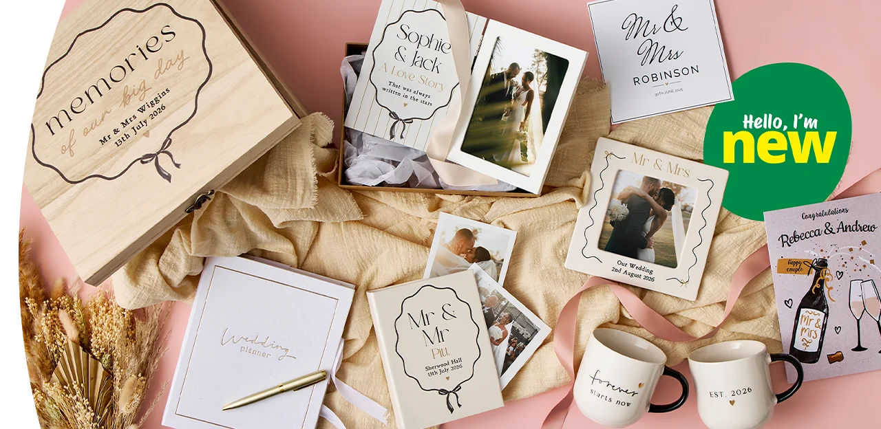 A flat lay of coordinated wedding gifts and keepsakes including a wooden memories box, personalised cards, photo prints, mugs, a notebook, ribbons and decorative foliage arranged on a soft pink background.