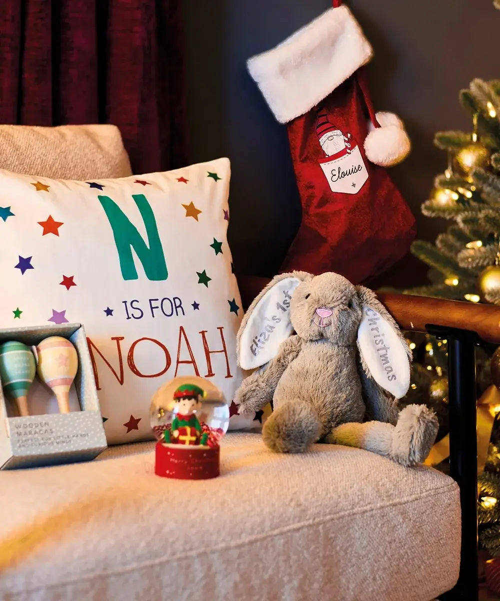 Cozy Christmas scene featuring a personalised pillow reading 'N is for NOAH', a red stocking labeled 'Louise', a plush bunny with 'My First Christmas' on its ears, wooden maracas, and a festive elf snow globe beside a decorated tree.