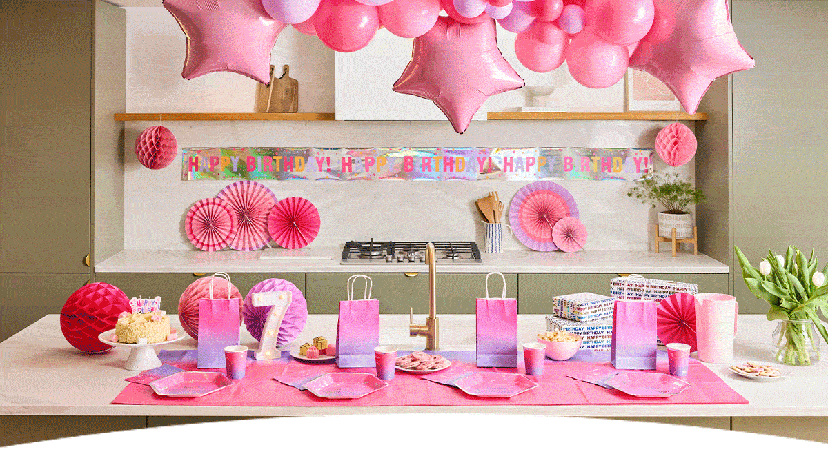 A pink-themed birthday party table decorated with a balloon arch, star-shaped foil balloons and a holographic ‘Happy Birthday!’ banner. The table is set with pink plates, cups, gift bags and party decorations, alongside a birthday cake, a number 7 candle, wrapped presents and a vase of white tulips in a modern kitchen setting.