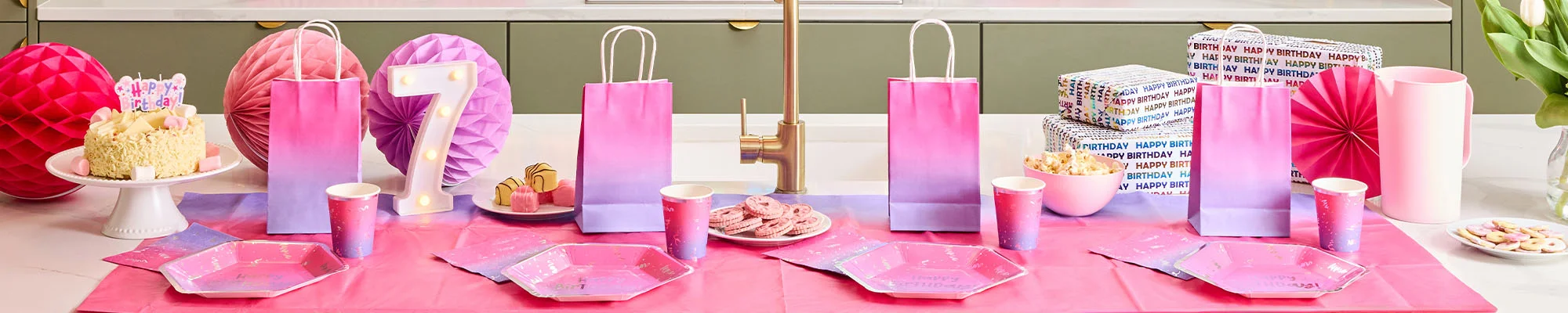A pink and purple ombré birthday party table display featuring matching party bags, plates and cups arranged on a pink table covering. The setup includes a birthday cake on a cake stand, a light-up number 7 decoration, wrapped presents, paper fan decorations, party food and snacks, creating a coordinated pastel birthday celebration theme. A pink and purple ombré birthday party table display featuring matching party bags, plates and cups arranged on a pink table covering. The setup includes a birthday cake on a cake stand, a light-up number 7 decoration, wrapped presents, paper fan decorations, party food and snacks, creating a coordinated pastel birthday celebration theme.