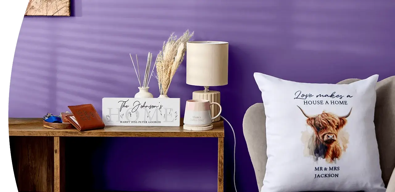A cosy home display featuring personalised wall art, a wooden clock, a table lamp, framed prints, decorative accessories and a cushion with a highland cow design arranged against a dark blue wall.