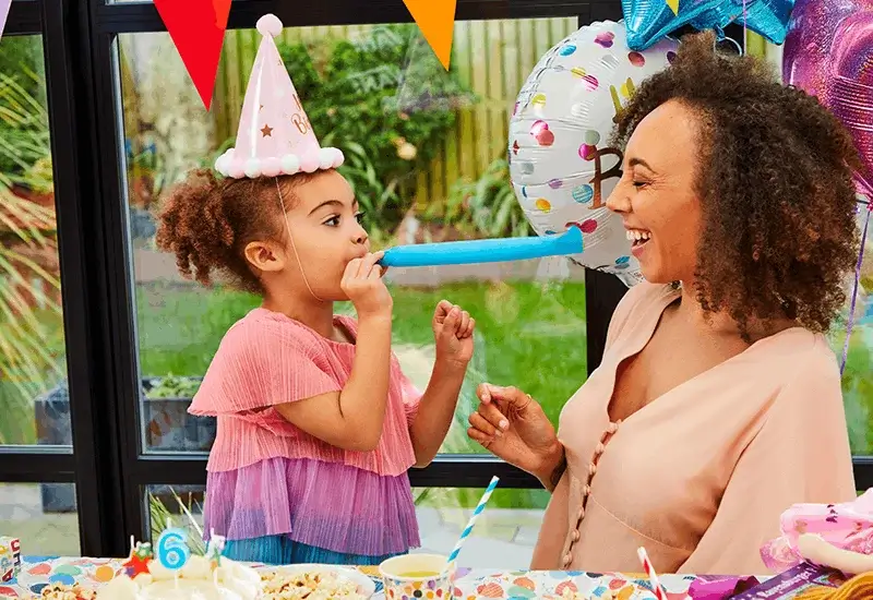 Child wearing a birthday hat blowing a party blower across a birthday table decorated with balloons, bunting, cake, and party tableware.